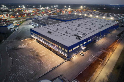 Aerial view of a large, illuminated industrial building next to a container depot at night